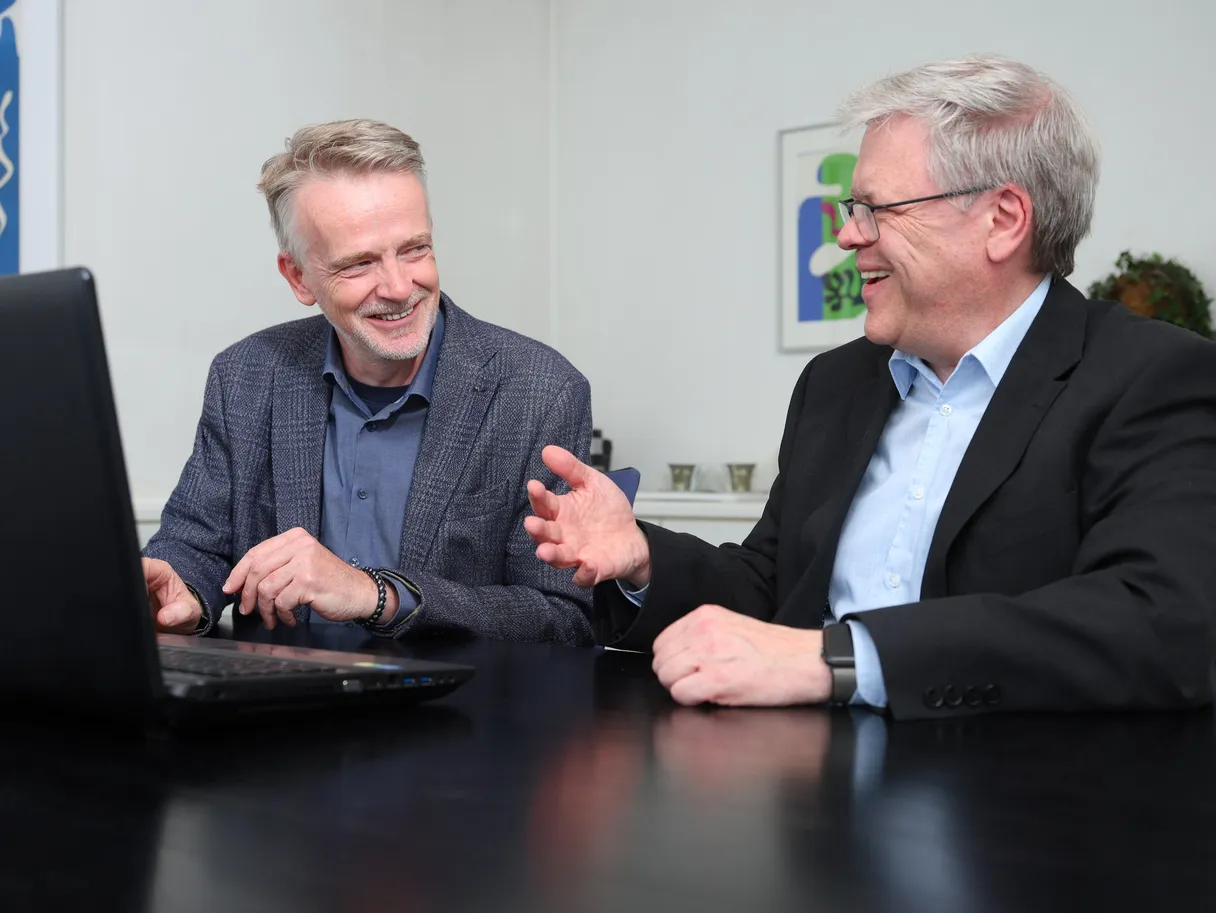 Die beiden Partner der Kanzlei sitzen lachend nebeneinander am Besprechungstisch vor einem Laptop.