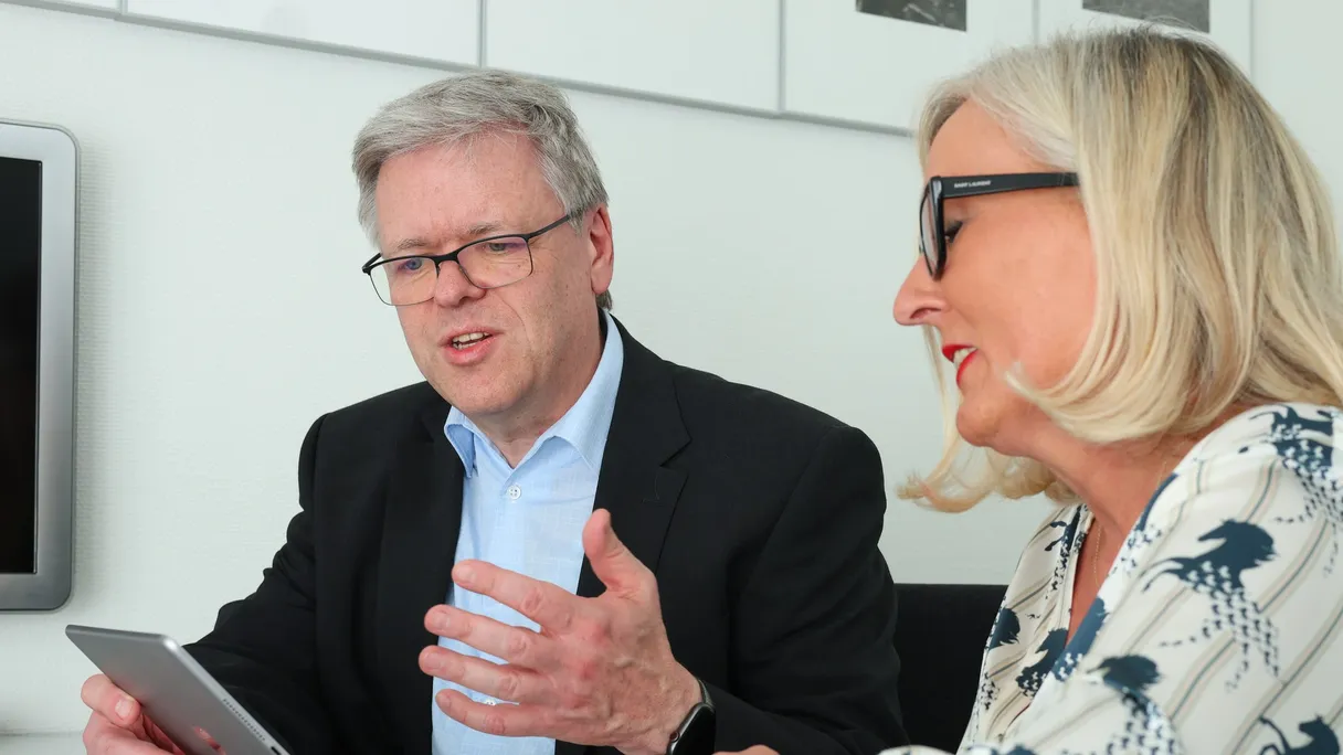 Ein Mann im schwarzen Sakko erklärt einer Frau mit Brille etwas, vor ihnen liegt ein Tablet.