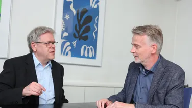 Die beiden Partner sitzen sich am Besprechungstisch gegenüber, zwischen ihnen Laptop und Gesetzbuch.