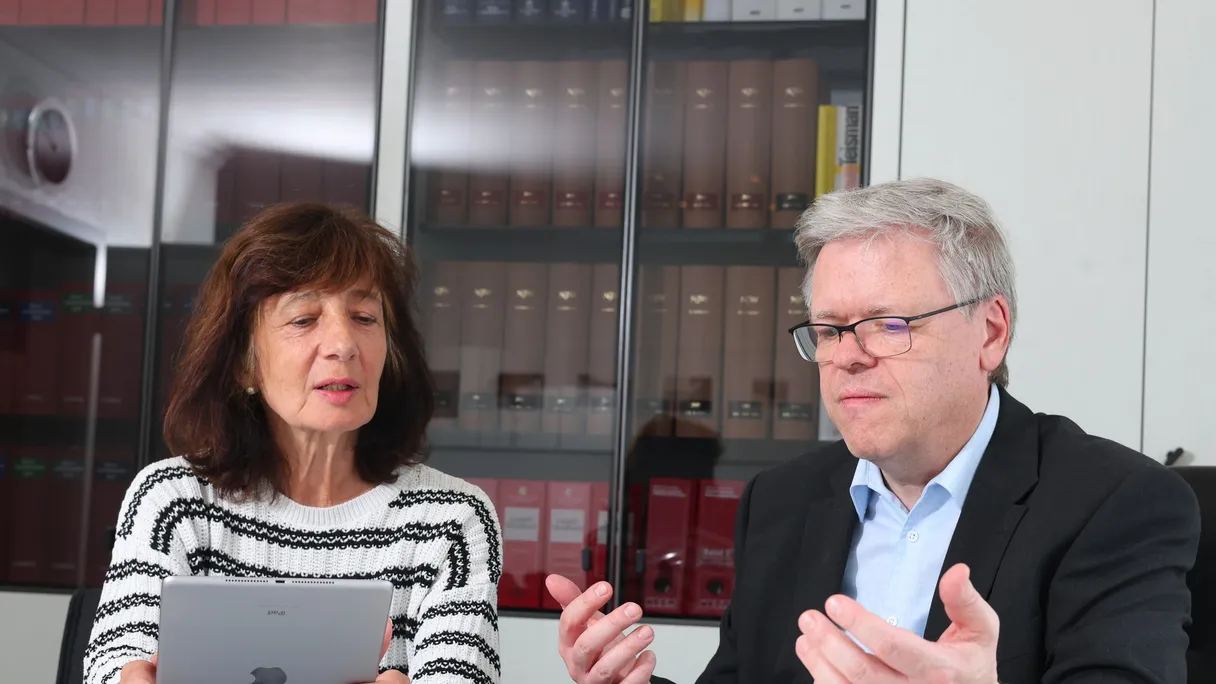 Ein Mann im schwarzen Sakko und eine Frau mit Tablet sitzen am Tisch, vor ihnen ein aufgeschlagenes Buch.