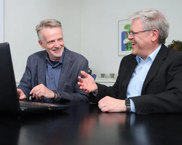 Die beiden Partner sitzen lachend nebeneinander am Besprechungstisch vor einem Laptop.