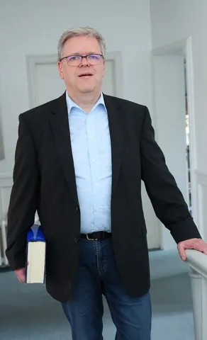 Ein Mann im schwarzen Sakko steht mit einem Buch unter dem Arm am Treppenabsatz.