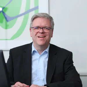 Porträt: Mann mit Brille im schwarzen Sakko und hellblauen Hemd, sitzend am Besprechungstisch.
