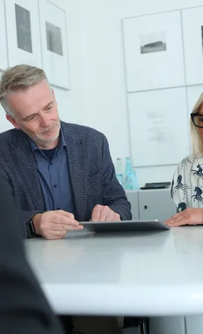 Ein Mann und eine Frau sitzen am runden Besprechungstisch und sehen auf ein Tablet.