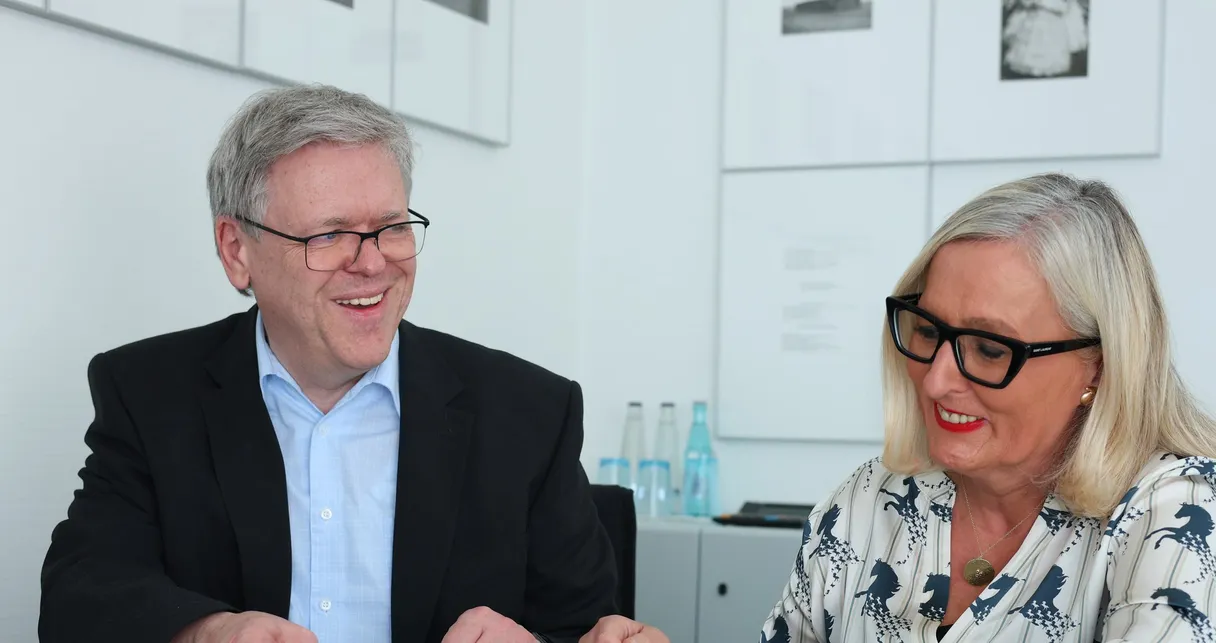 Ein Mann im schwarzen Sakko und eine Frau mit Brille sitzen lachend am runden Besprechungstisch, vor ihnen ein Tablet.