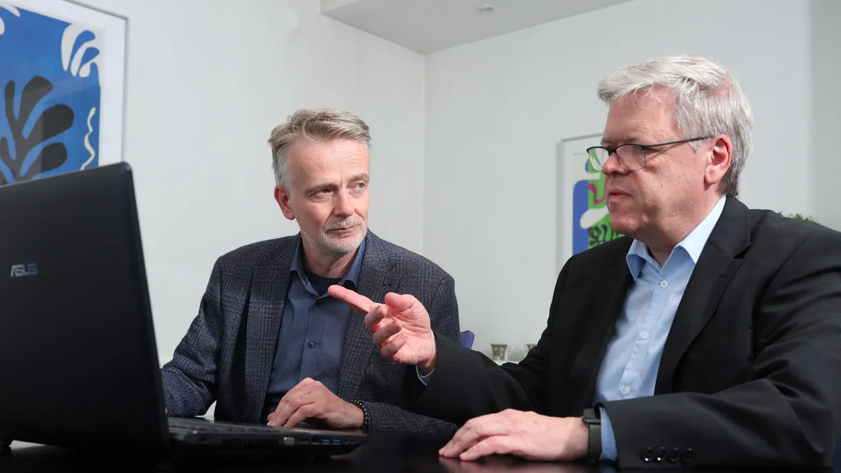 Die beiden Partner sitzen am Besprechungstisch vor einem Laptop und sprechen miteinander.