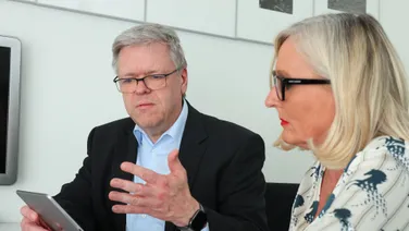 Ein Mann im schwarzen Sakko und eine Frau mit Brille sprechen am Besprechungstisch über ein Tablet.