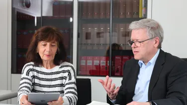 Eine Frau mit Tablet und ein Mann im schwarzen Sakko sitzen am Tisch vor einem aufgeschlagenen Buch.