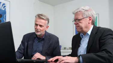 Die beiden Partner sitzen nebeneinander am Besprechungstisch vor einem Laptop.