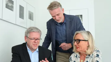 Ein Mann steht hinter zwei sitzenden Personen, alle drei sehen auf ein Tablet.