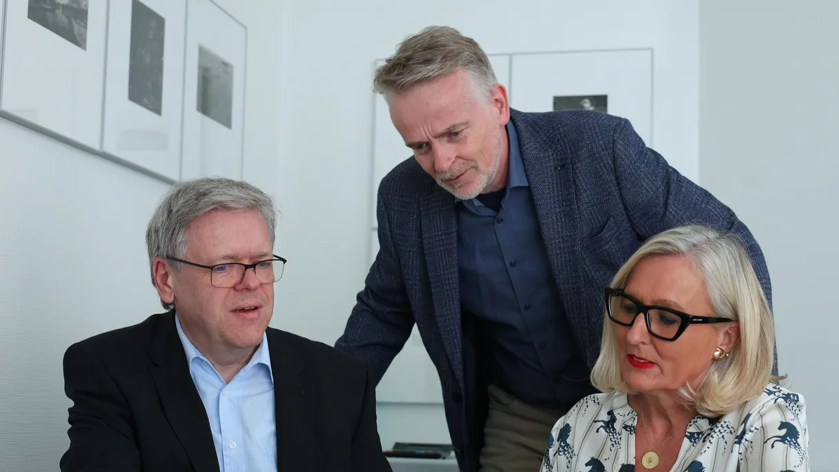 Ein Mann steht hinter zwei sitzenden Personen und alle drei sehen auf einen Laptop.